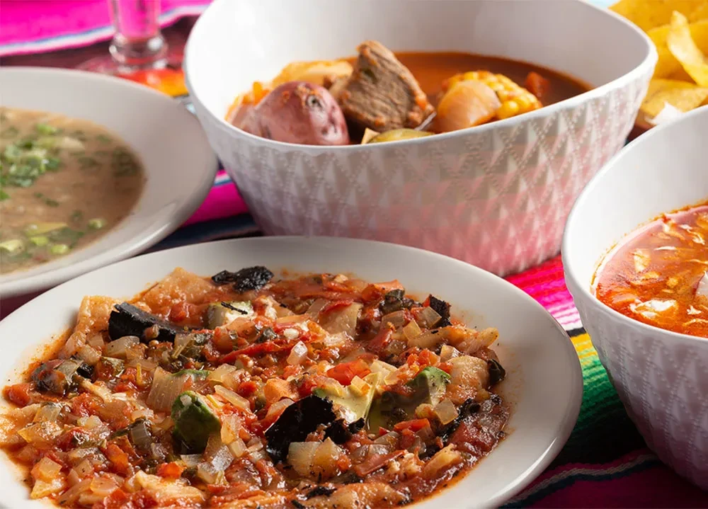 Vibrant Mexican meal spread with stews, soups on colorful serape.