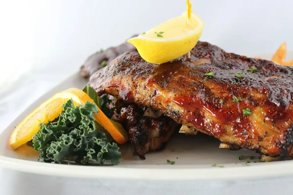 Close-up of barbecued ribs with glossy sauce, lemon, kale, and orange.