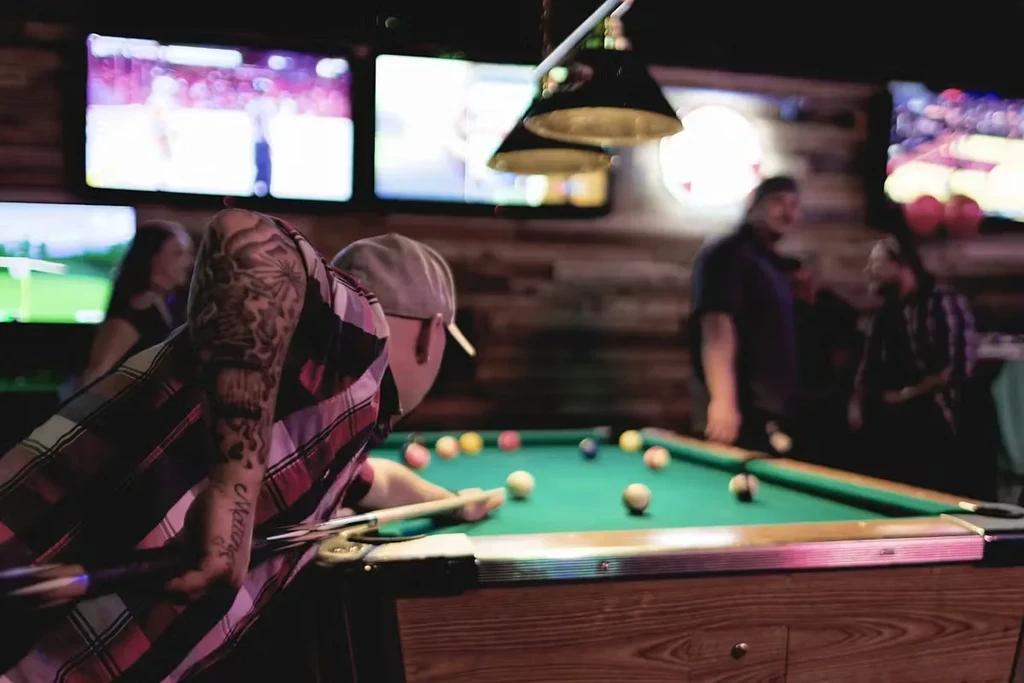 Tattooed man plays pool in a dim, busy bar.