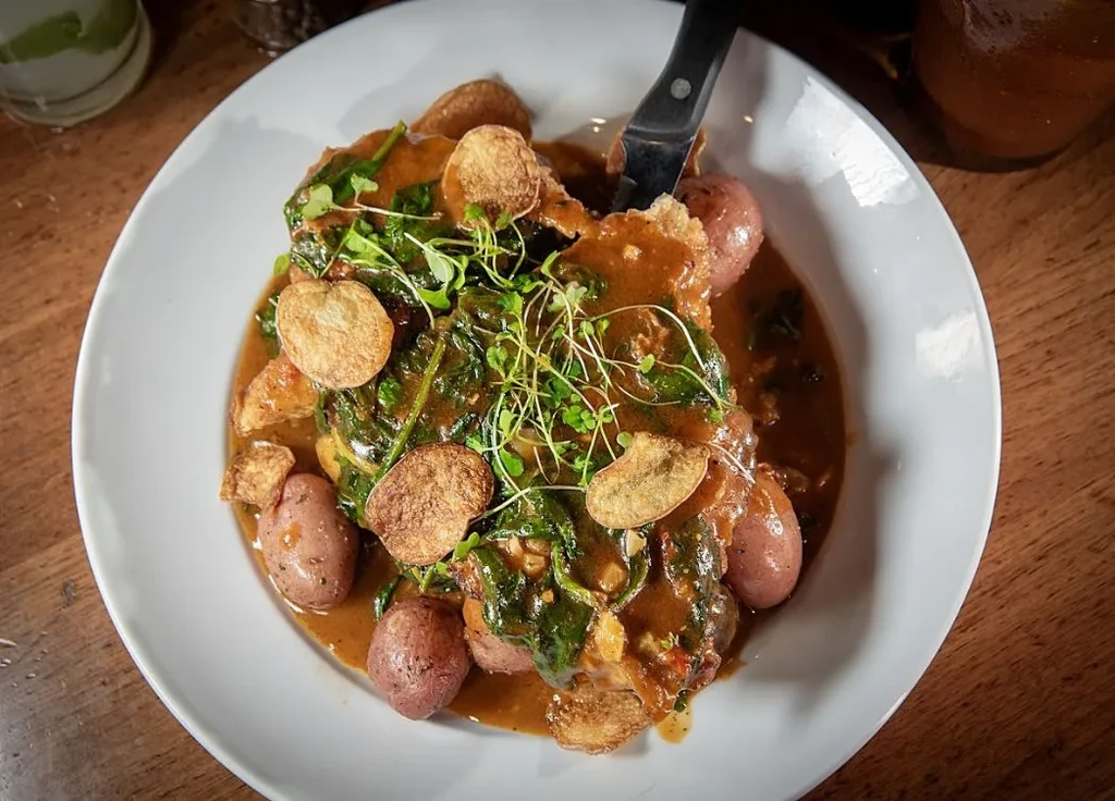 Potatoes, greens, and sauce in a white bowl with a knife.