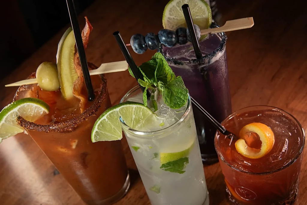 Four distinctly garnished cocktails with straws on a dark wooden surface.