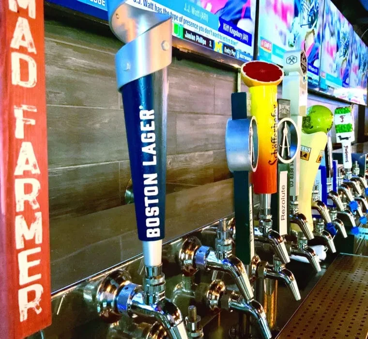 Close-up of diverse beer taps with Boston Lager prominent, sports TVs above.