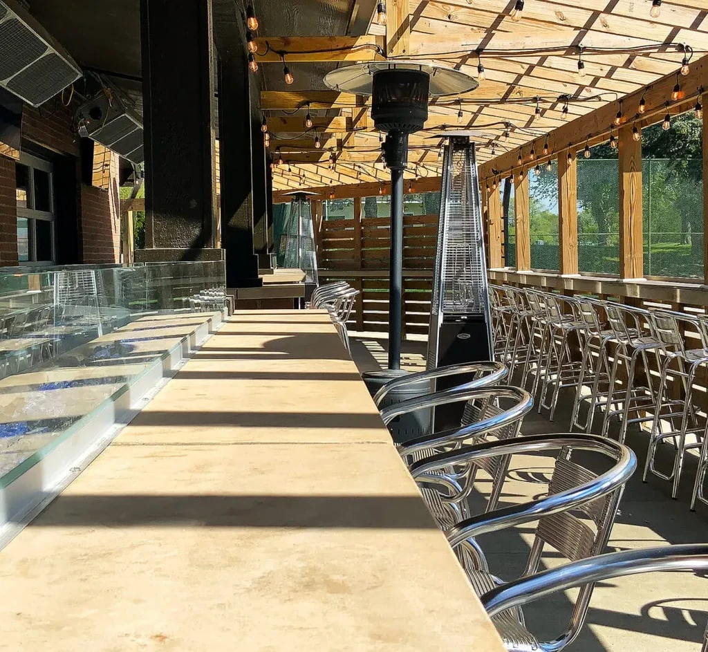 Outdoor patio bar with silver stools, trellis, string lights, and heaters.