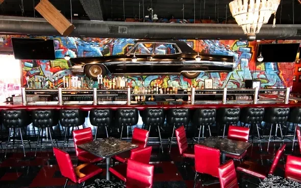 Vibrant bar interior with red seating, graffiti, and vintage car on wall.