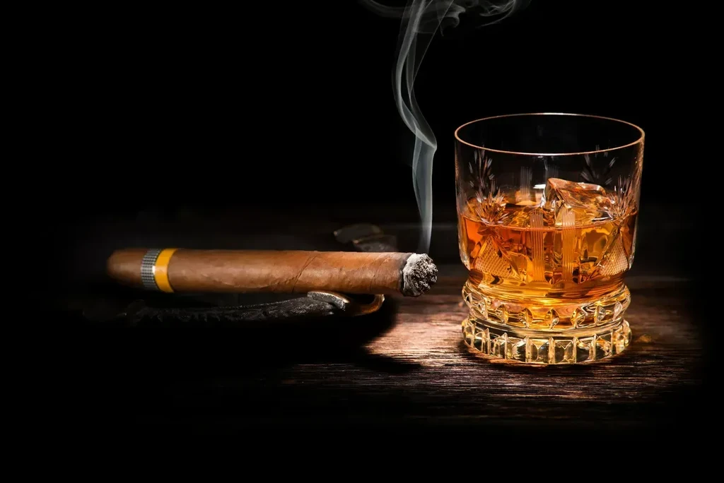 Lit cigar on ashtray next to whiskey glass, smoke rising.