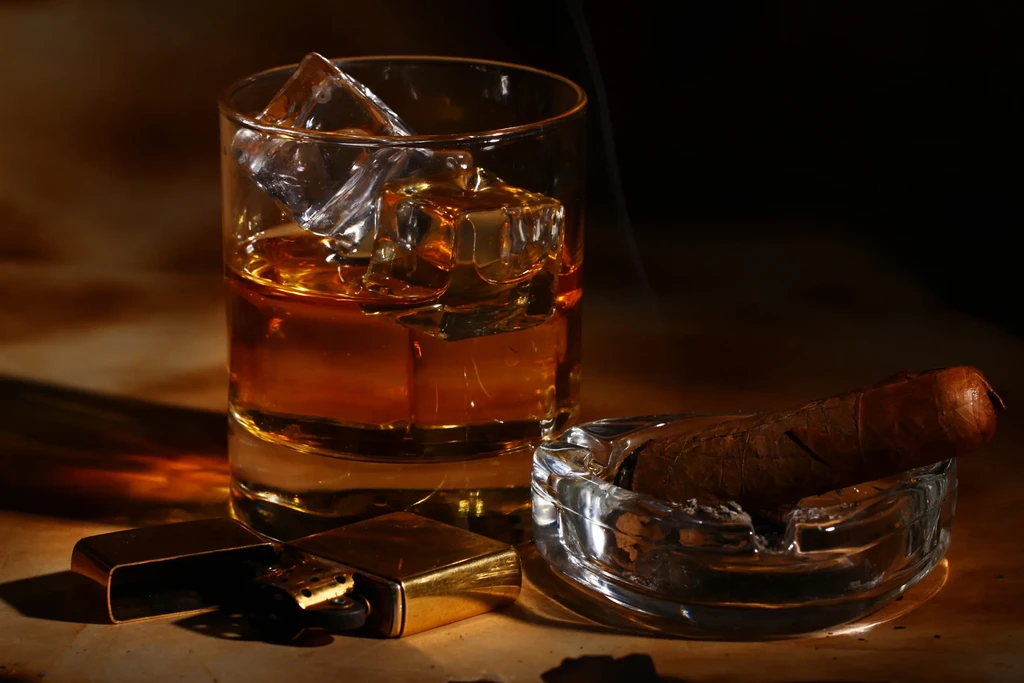 Dimly lit close-up of whiskey with ice, lit cigar, and open lighter.