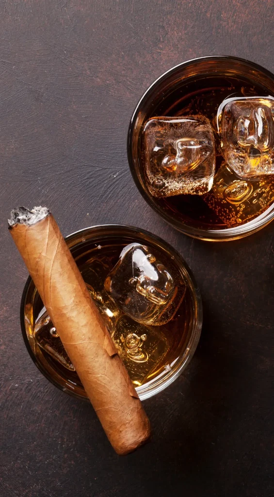 Top-down: Two amber drinks with ice; one has a lit cigar.