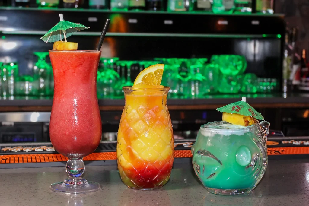 Three colorful tropical drinks with garnishes on a green-lit bar.