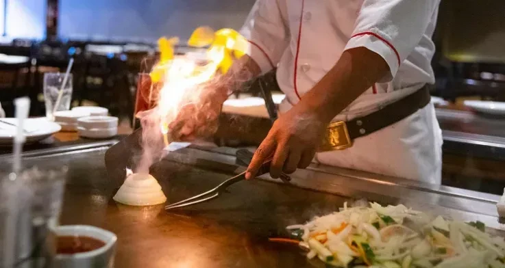 Japanese hibachi chef cooks on grill, flaming onion volcano erupts.
