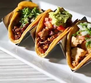 Three tacos with diverse toppings on a white rectangular plate.