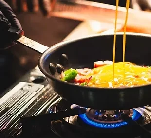 Gloved hand pours eggs into pan with food over gas flame.
