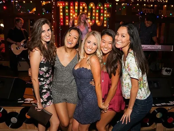Five women pose in a bar with a band and "TIN ROOF" sign.