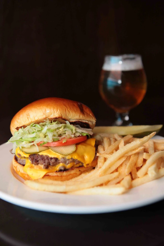 Double cheeseburger, fries, pickle, and beer served on a white plate.