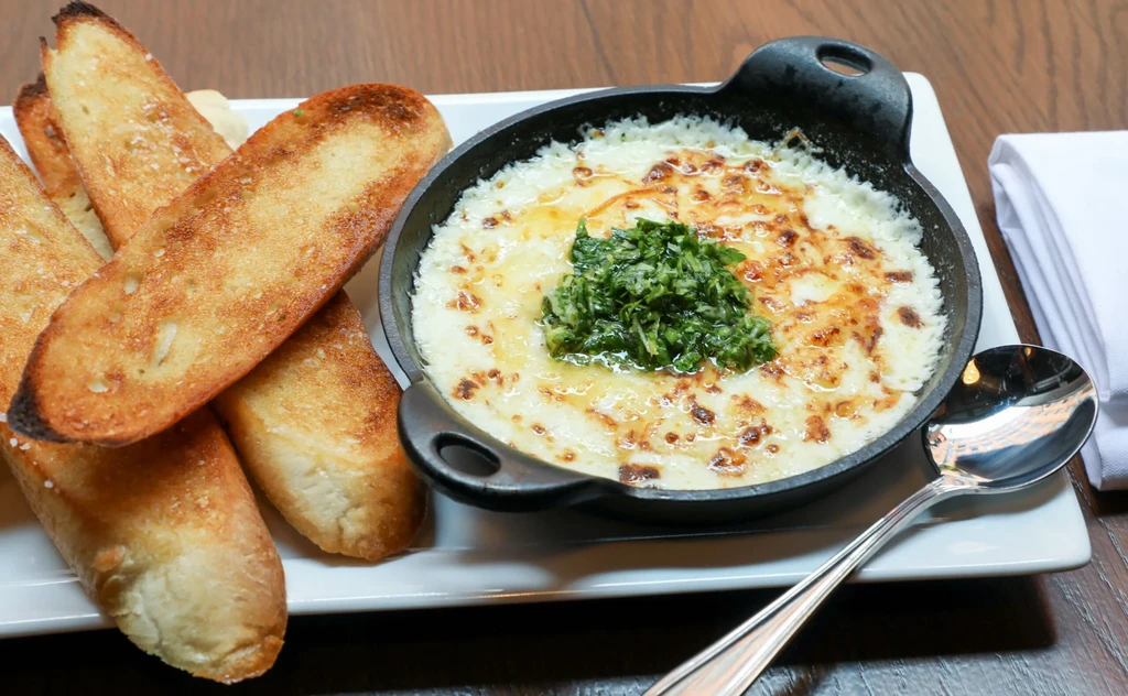 Skillet of bubbly cheese dip with herbs and toasted baguette.