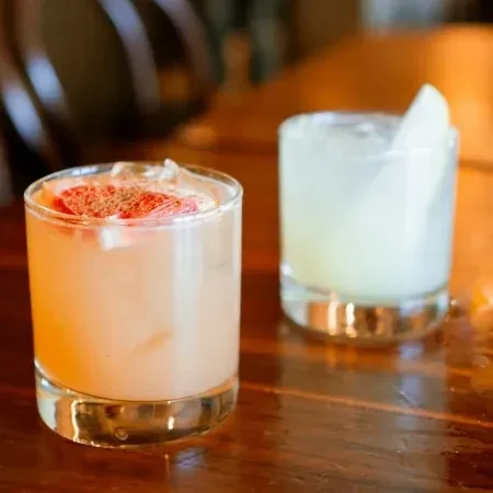 Two short cocktails on a wooden bar, garnished with citrus and fruit.