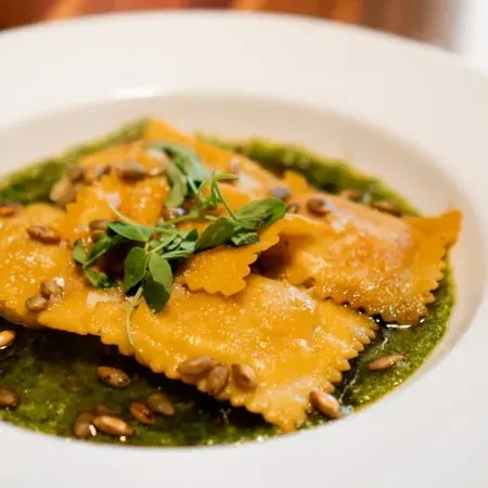 Orange ravioli in green sauce with pumpkin seeds and herb garnish.