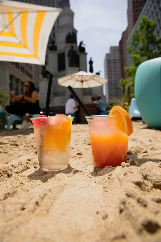 Two colorful layered drinks on sand, blurred city background.
