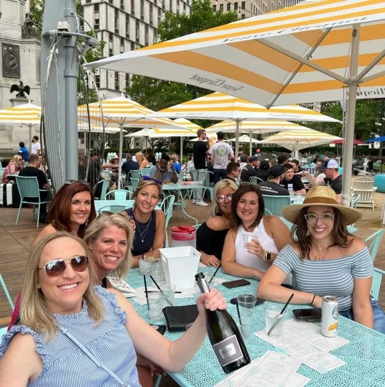 Seven smiling women at an outdoor bar with an Avissi bottle.