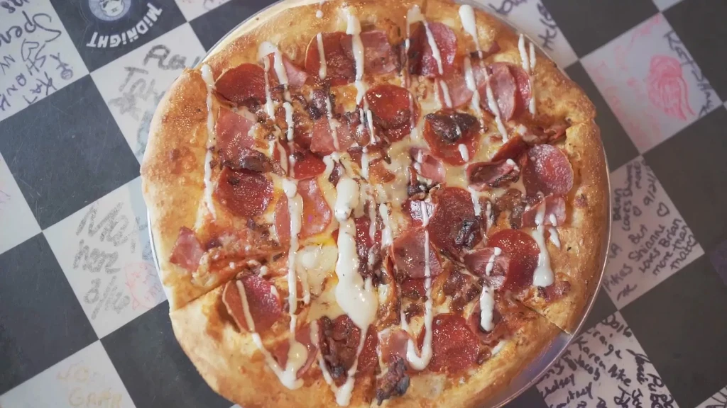 Round meat pizza with white drizzle, served on a signed checkered table.