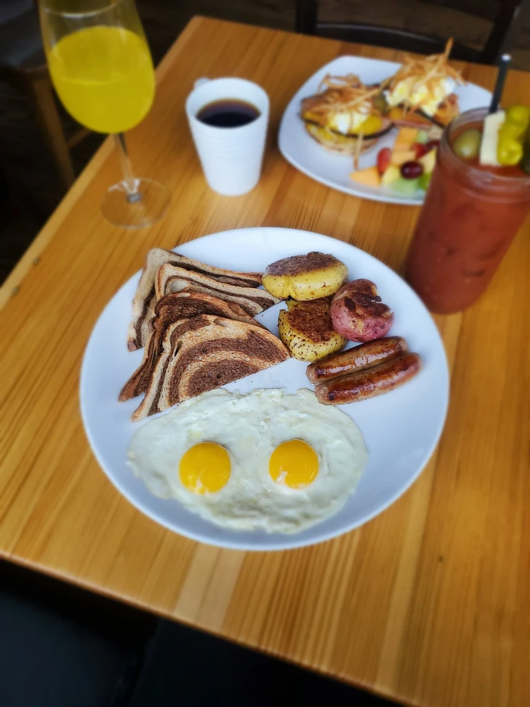 Breakfast spread with fried eggs, rye toast, sausages, potatoes, and drinks.