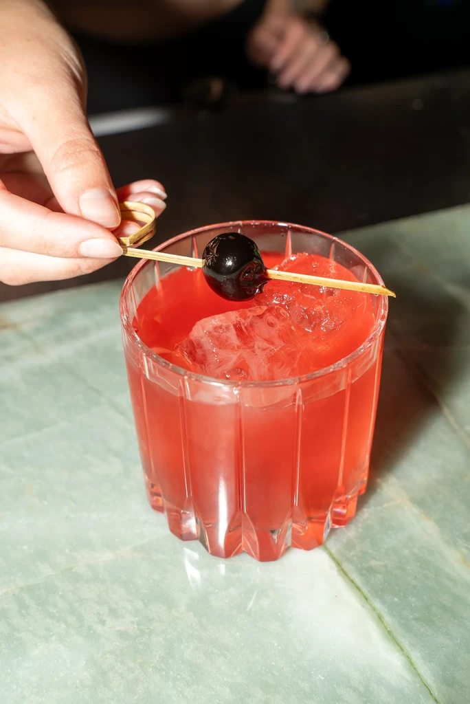 Hand garnishing a pink cocktail with a cherry and crushed ice.