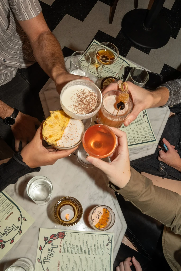 Top-down: Diverse hands clink colorful cocktails on a white marble table.