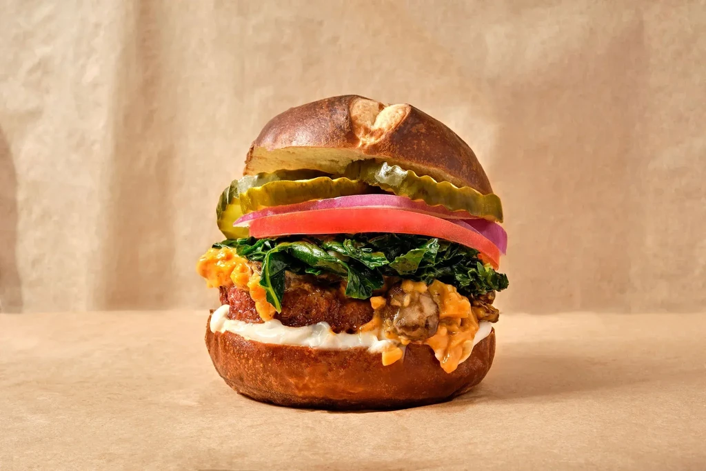 Gourmet burger with dark bun, thick patty, greens, tomato, and pickles.