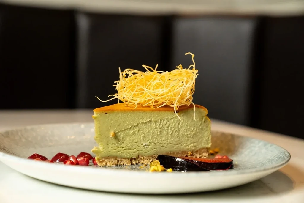 Slice of pale green dessert, kataifi, pomegranate, and dark fruit on a plate.