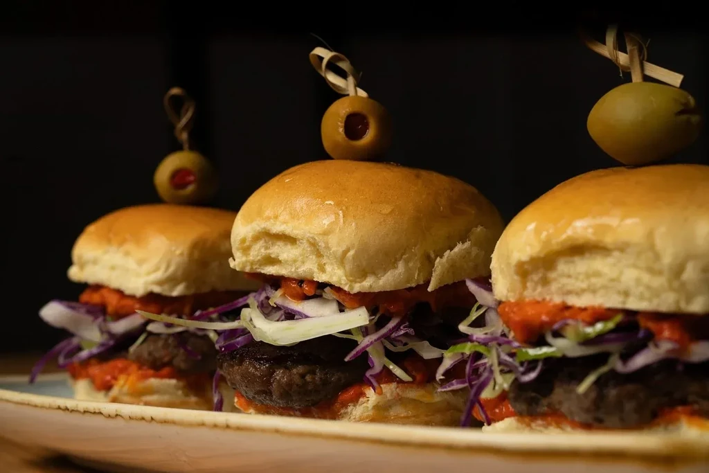 Three sliders with buns, meat, red sauce, slaw, and olive picks.