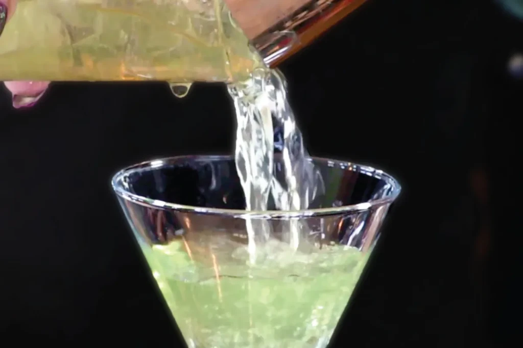 Hand pours bubbling green liquid into a martini glass.