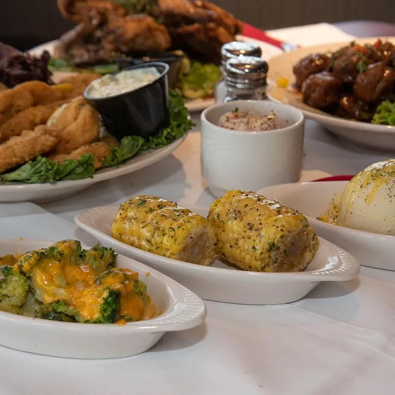 Dining table spread with cheesy broccoli, corn, mashed potatoes, and entrees.