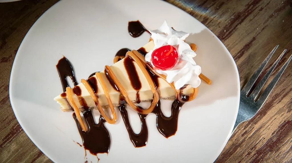 Cheesecake slice with chocolate, caramel, whipped cream, and cherry.