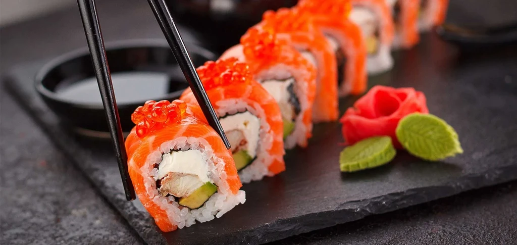 Chopsticks lift salmon, avocado, and roe sushi from a dark tray.