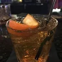 Golden cocktail in textured glass, garnished with ice, orange, and straw.