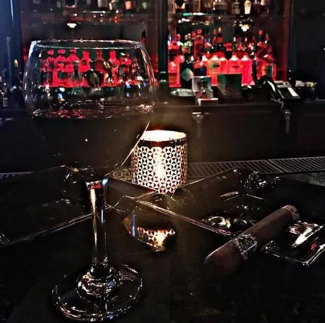 Dimly lit bar with wine glass, glowing candle, cigar, and colorful bottles.