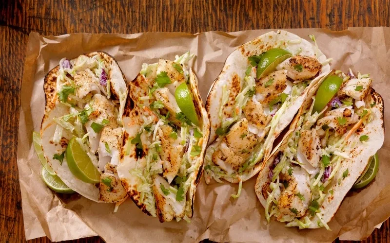 Four fish tacos with slaw, cilantro, and lime on parchment paper.