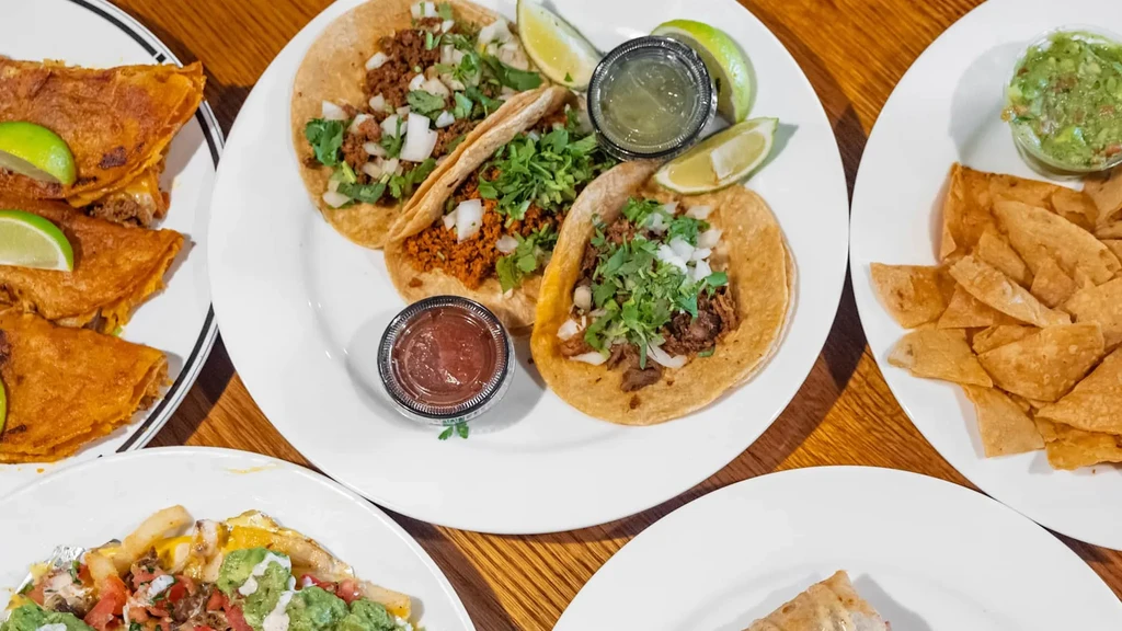 Overhead view of various Mexican dishes, including tacos, on wood table.