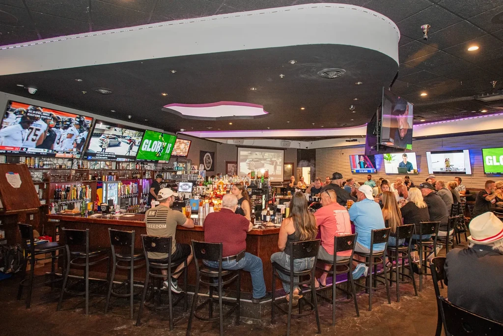 Busy modern sports bar with patrons, TVs, and purple accent lighting.