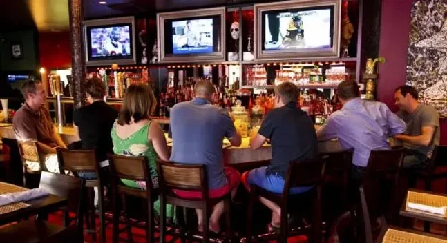 Seven patrons at a lively, dim bar with sports on TVs.