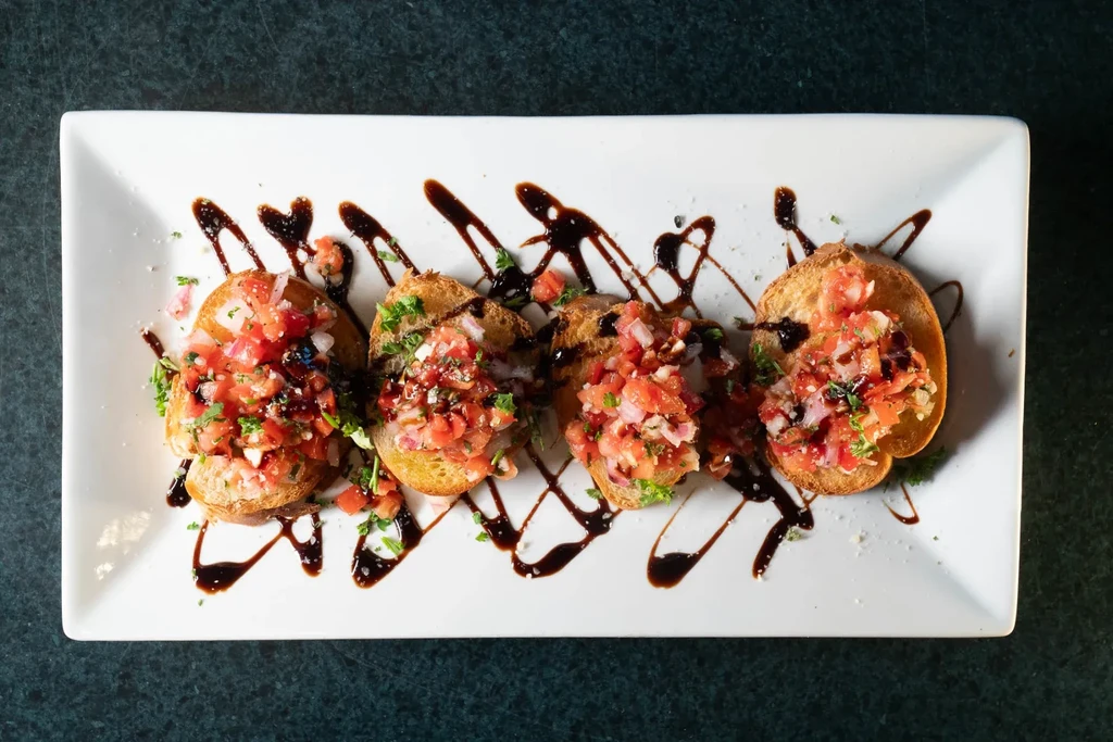 White plate with four tomato bruschetta, herbs, and balsamic glaze.
