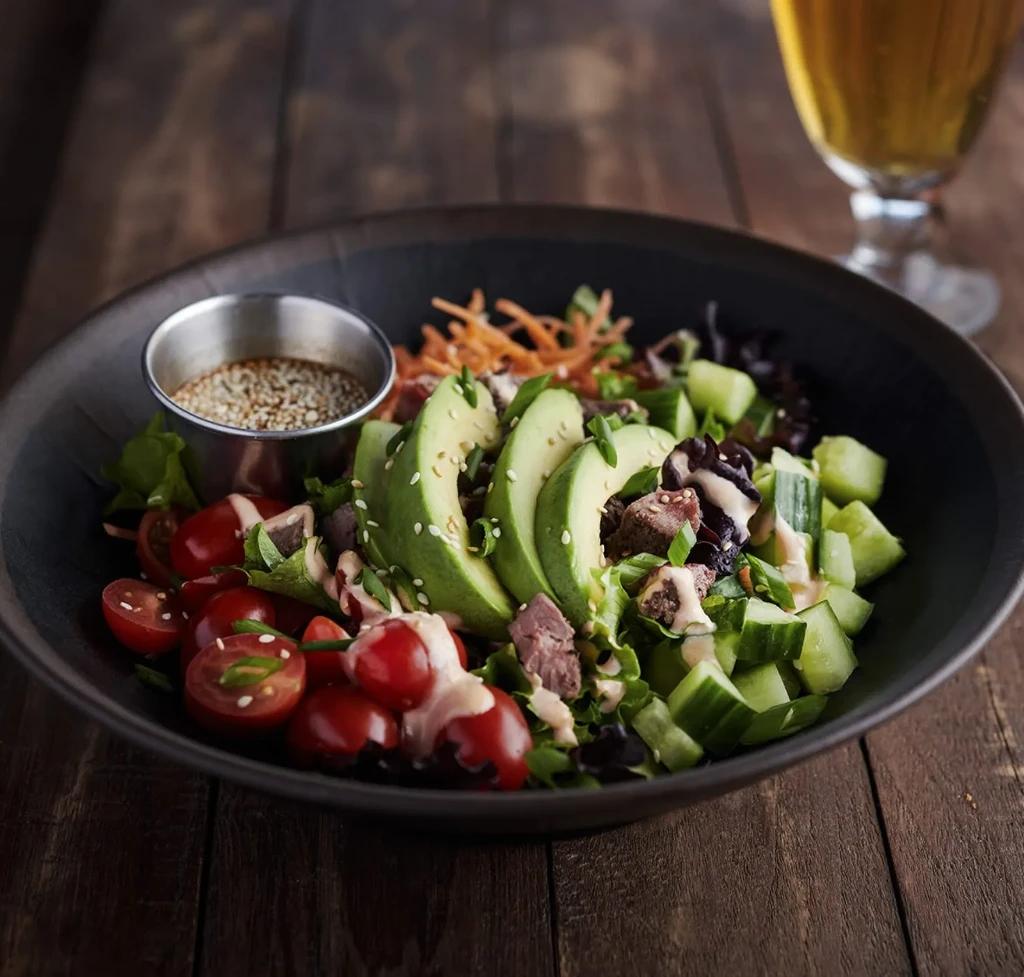 Salad with avocado, tomatoes, meat, and dressing in a dark bowl.