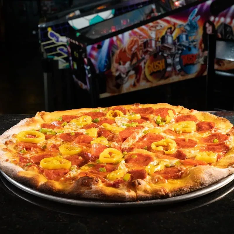 Pepperoni pizza with peppers on tray next to RUSH pinball machine.