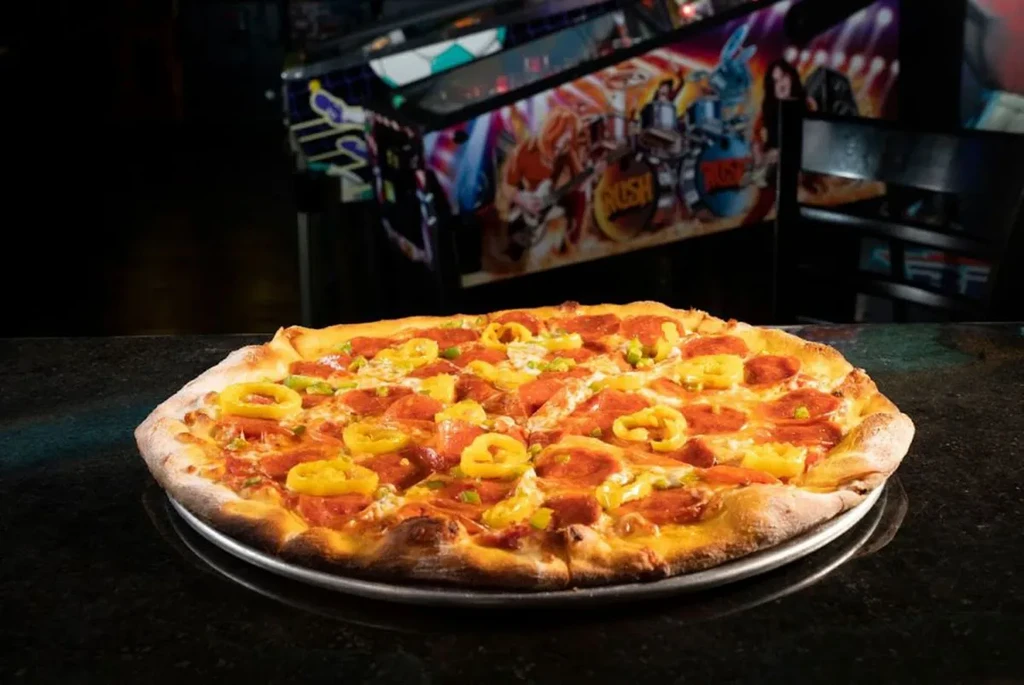 Pepperoni pizza with peppers on a pan, by a pinball machine.