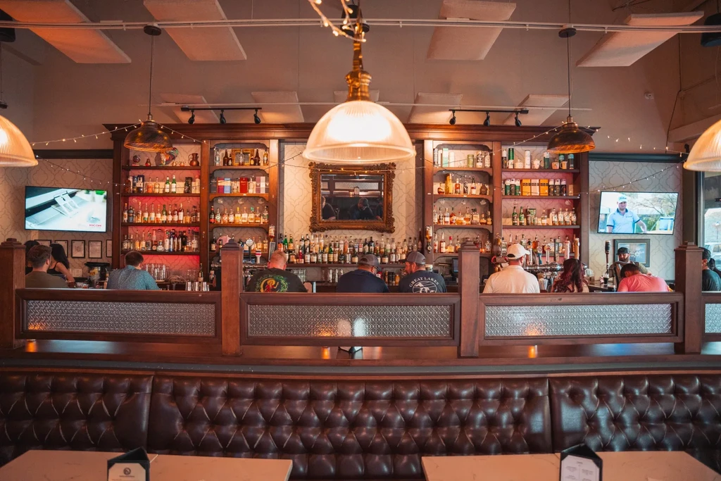 Ornate, well-lit bar interior with bottles, patrons, and tufted leather booths.