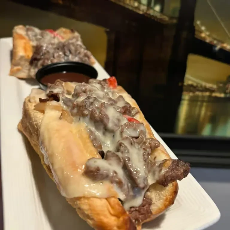 Two loaded cheesesteak subs on a plate, with dipping sauce.