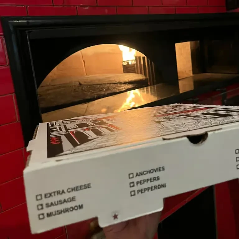 Pizza box with topping checklist held before a glowing wood-fired oven.