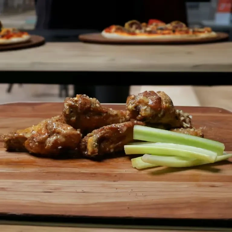 Golden-brown chicken wings and celery on a board, with pizzas.