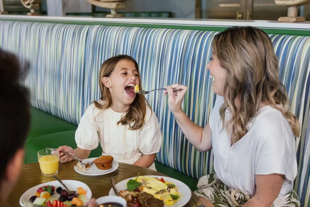 Woman feeding a laughing girl breakfast at a restaurant booth.