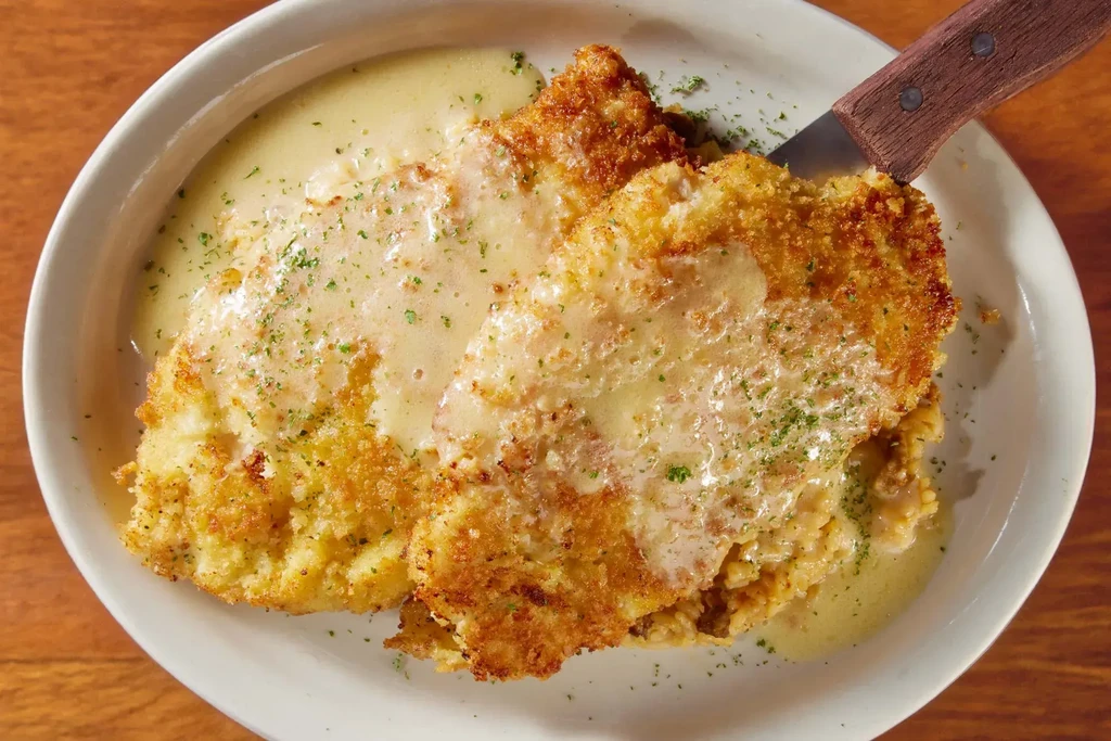 Two breaded cutlets with creamy gravy, chopped green herbs, knife inserted.