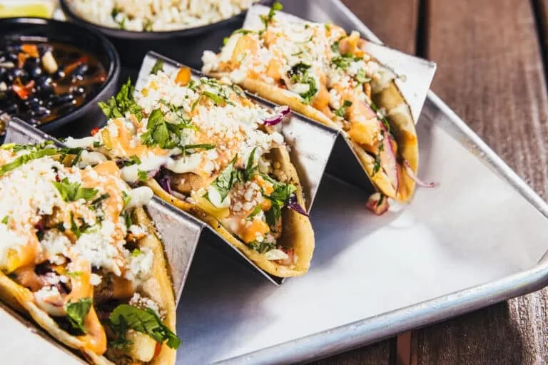 Four generously filled tacos in a metal holder on a tray.